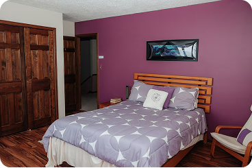 Bedroom with wooden bed frame, purple patterned bedding, wooden closet doors, and a purple accent wall with a framed picture.
