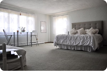 Cozy bedroom with a tufted headboard bed, white bedding and pillows, a small round table with chairs, and light coming through sheer curtains.