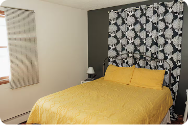 Bedroom with a bed covered in yellow bedding, a small bedside table with a lamp, and window with blinds.