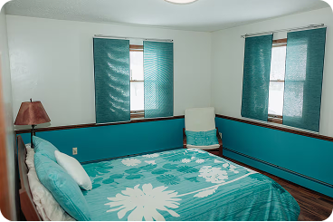 Bedroom with teal floral bedspread, teal curtains on windows, and a white armchair with a teal cushion in the corner.