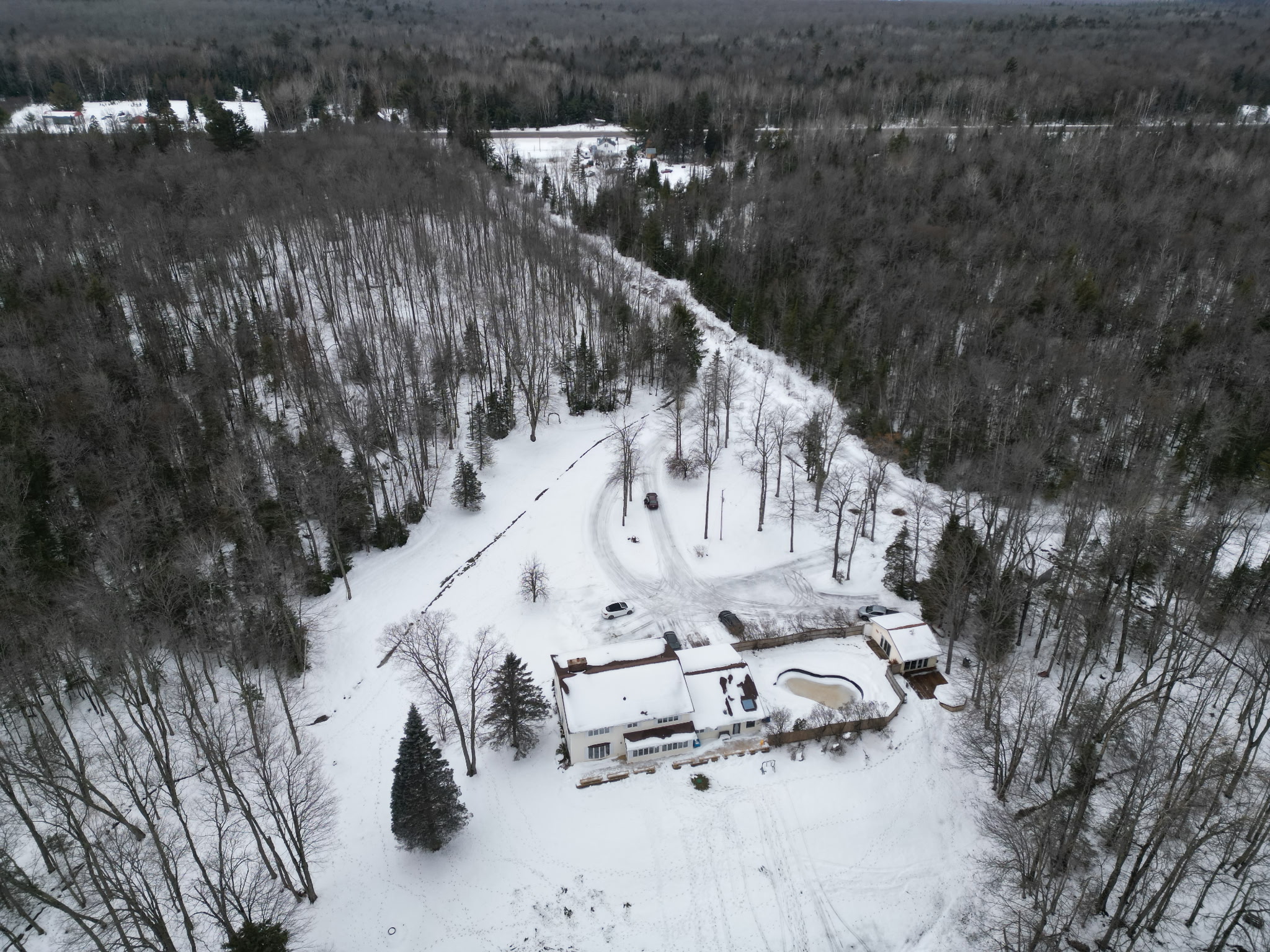 Aerial view of a snow-covered house with a fenced pool, surrounded by leafless trees and a snowy forest landscape.