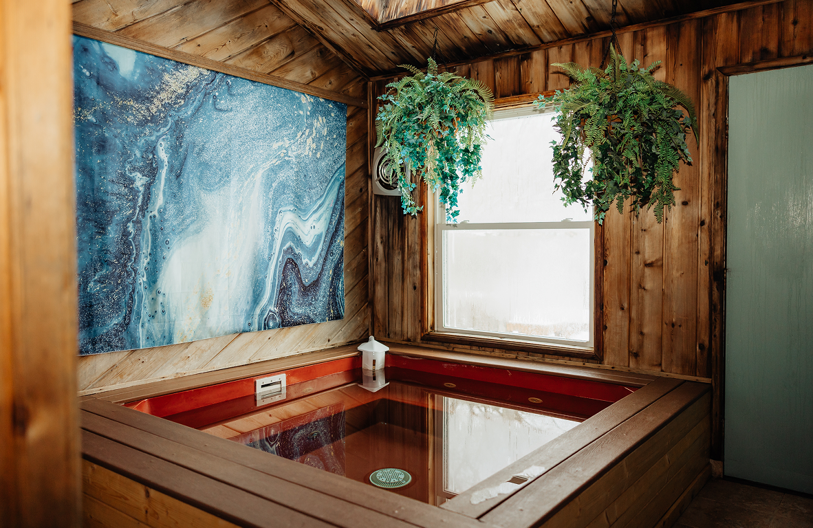 Wood-paneled room with a square hot tub, two hanging green plants, a large abstract blue wall art, and a window with frosted glass.