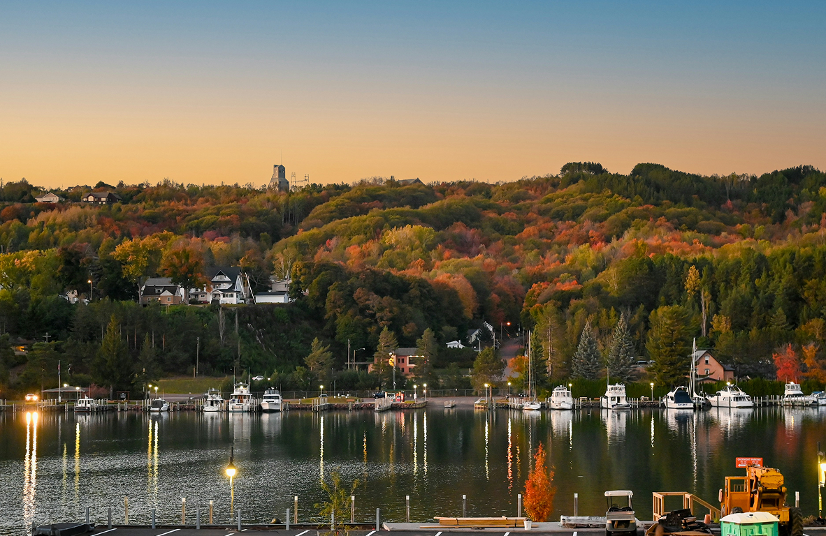 Calm lake reflecting lights and boats with a densely forested hillside showing autumn colors and houses under a clear sky at dusk.
