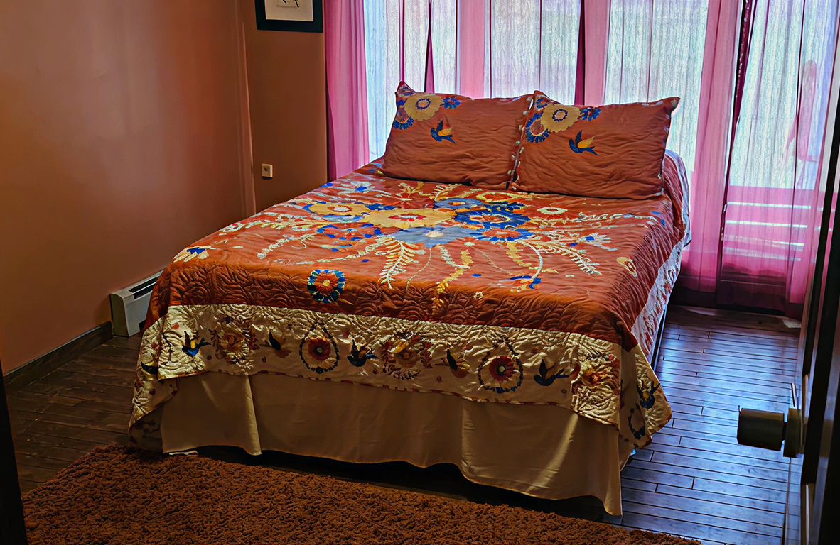 Bed with orange floral embroidered bedspread and matching pillows in a room with pink curtains and wooden floor.