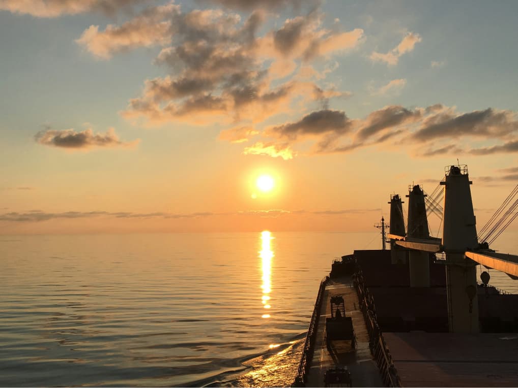 A scenic photograph of a sunset out at sea, taken from a large boat, the boats front in view in the right of the photo