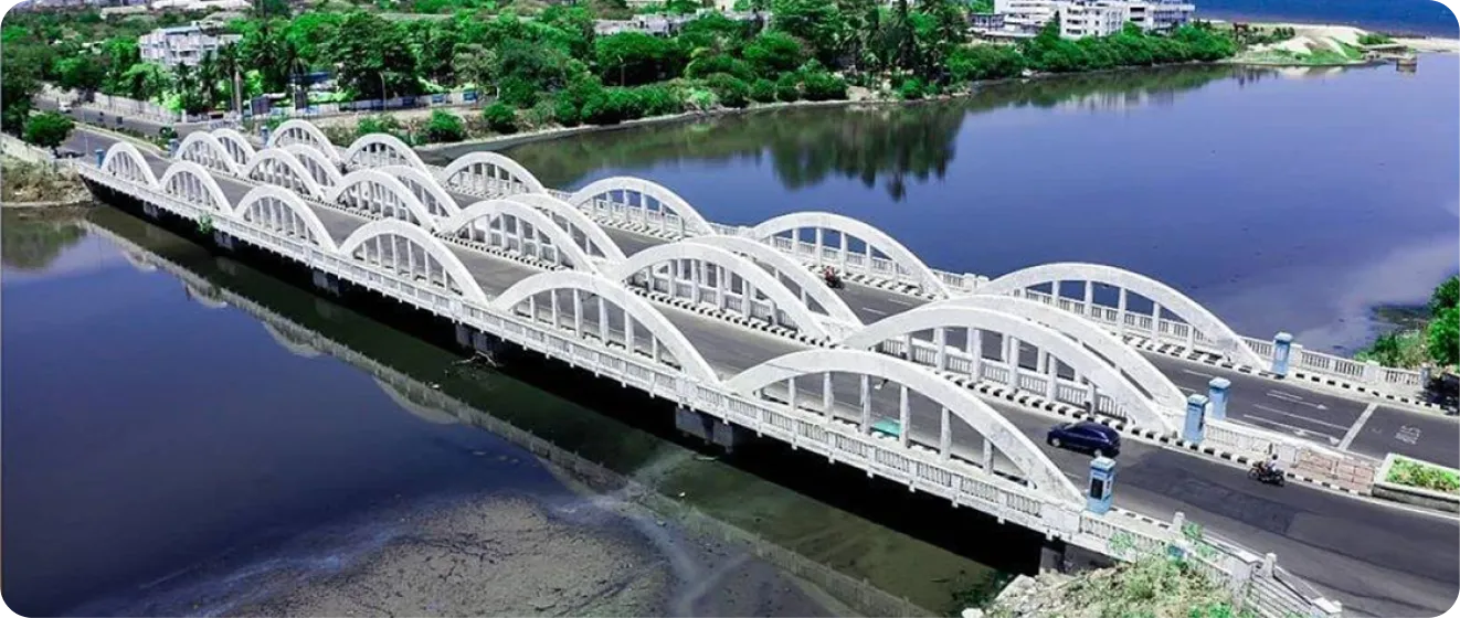 Wide white arched bridge over calm river with vehicles and greenery in the background.