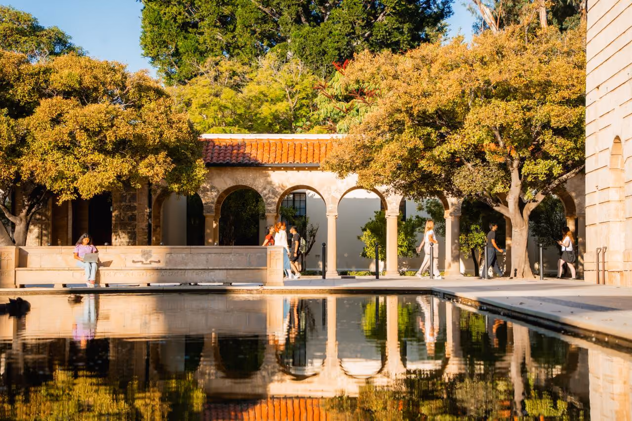 Sunny outdoor courtyard with arched stone colonnade, trees with autumn foliage, and people walking by a reflective rectangular pond.
