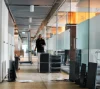 Modern office corridor with glass partitions, desks, chairs, and a person walking in the distance.