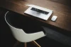 Modern white chair with wooden legs next to a dark wooden desk holding an open laptop, smartphone, and notebook.
