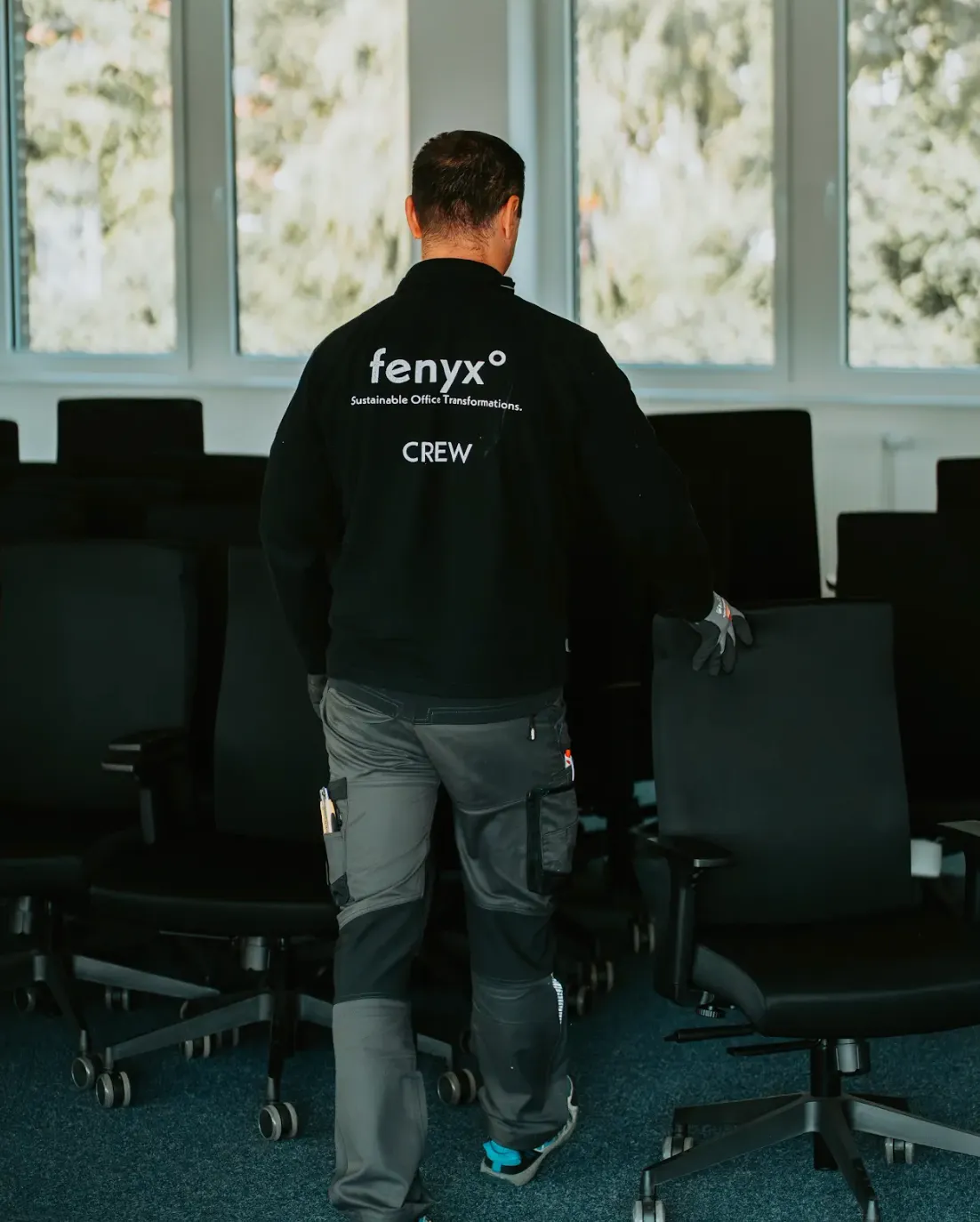 Person wearing a black jacket labeled 'fenyx Sustainable Office Transformations CREW' pushing a black office chair in a room with several chairs and large windows.