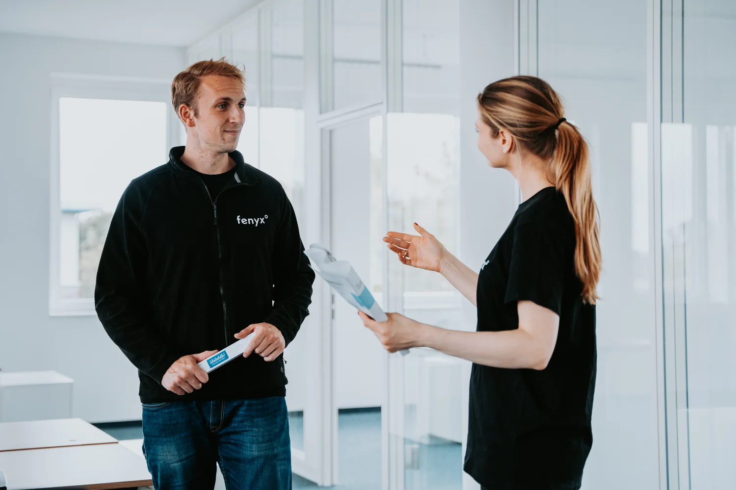 Two coworkers in an office talking, one holding paperwork and the other holding a small object.