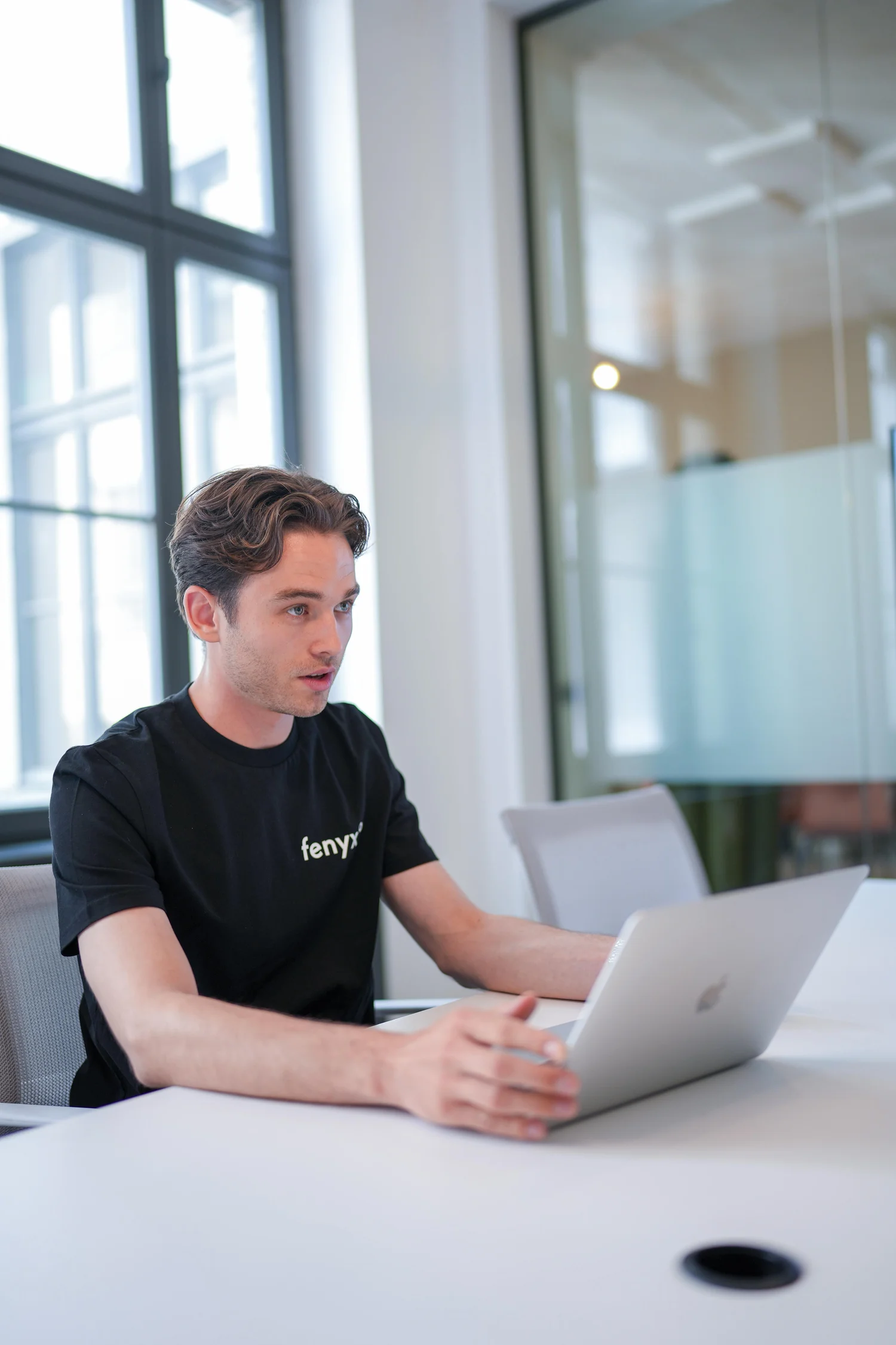 Young man wearing a black fenyx t-shirt working on a silver laptop at a white office table.