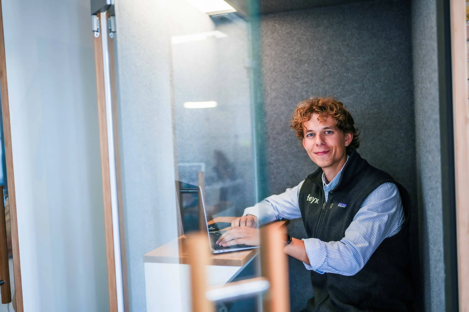 Young man with curly hair smiling while working on a laptop in a small modern office booth.