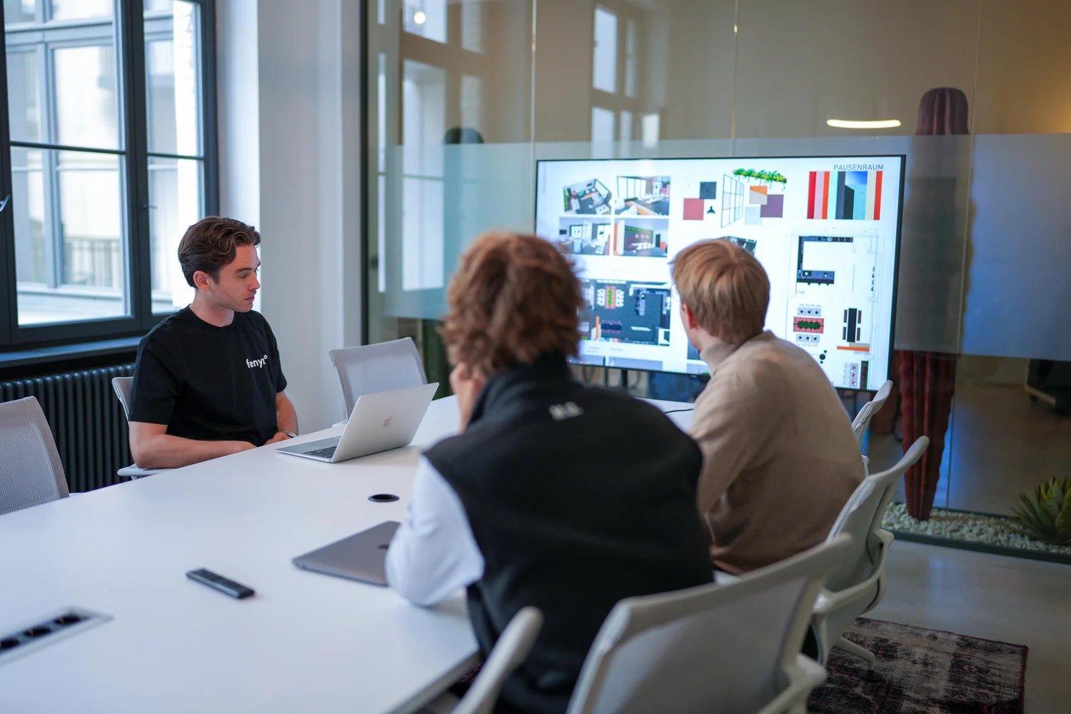 Three men in a modern conference room viewing architectural or design plans displayed on a large screen.