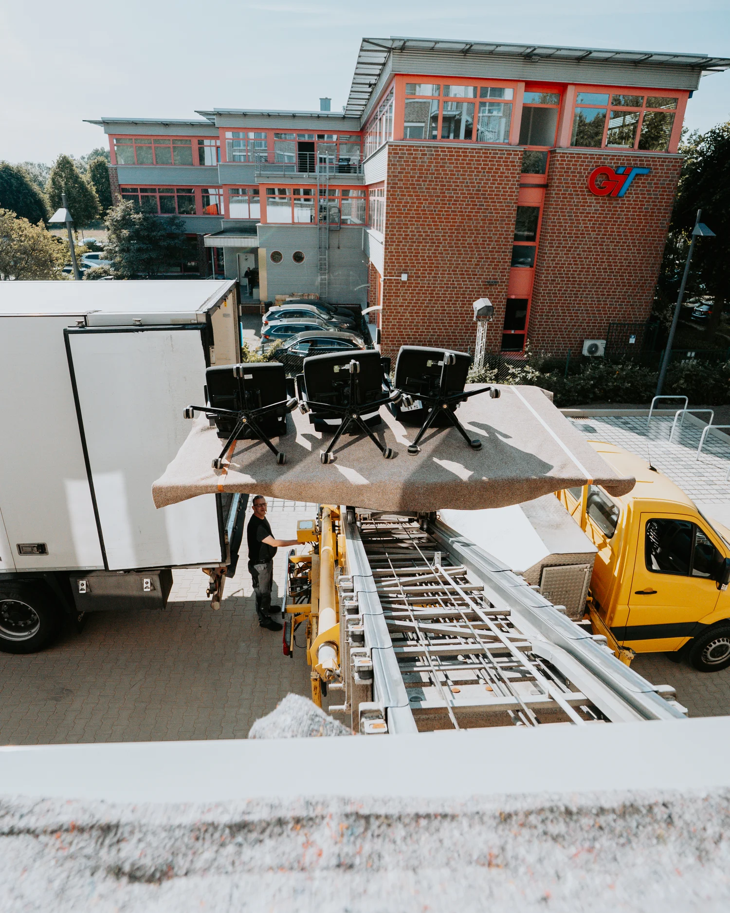 Three office chairs being lifted on a platform by a yellow crane truck near a brick building with the GT logo.