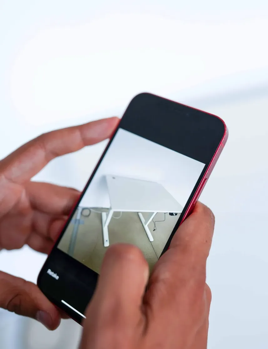 Person holding a smartphone displaying a photo of a white desk with angled legs against a white wall.