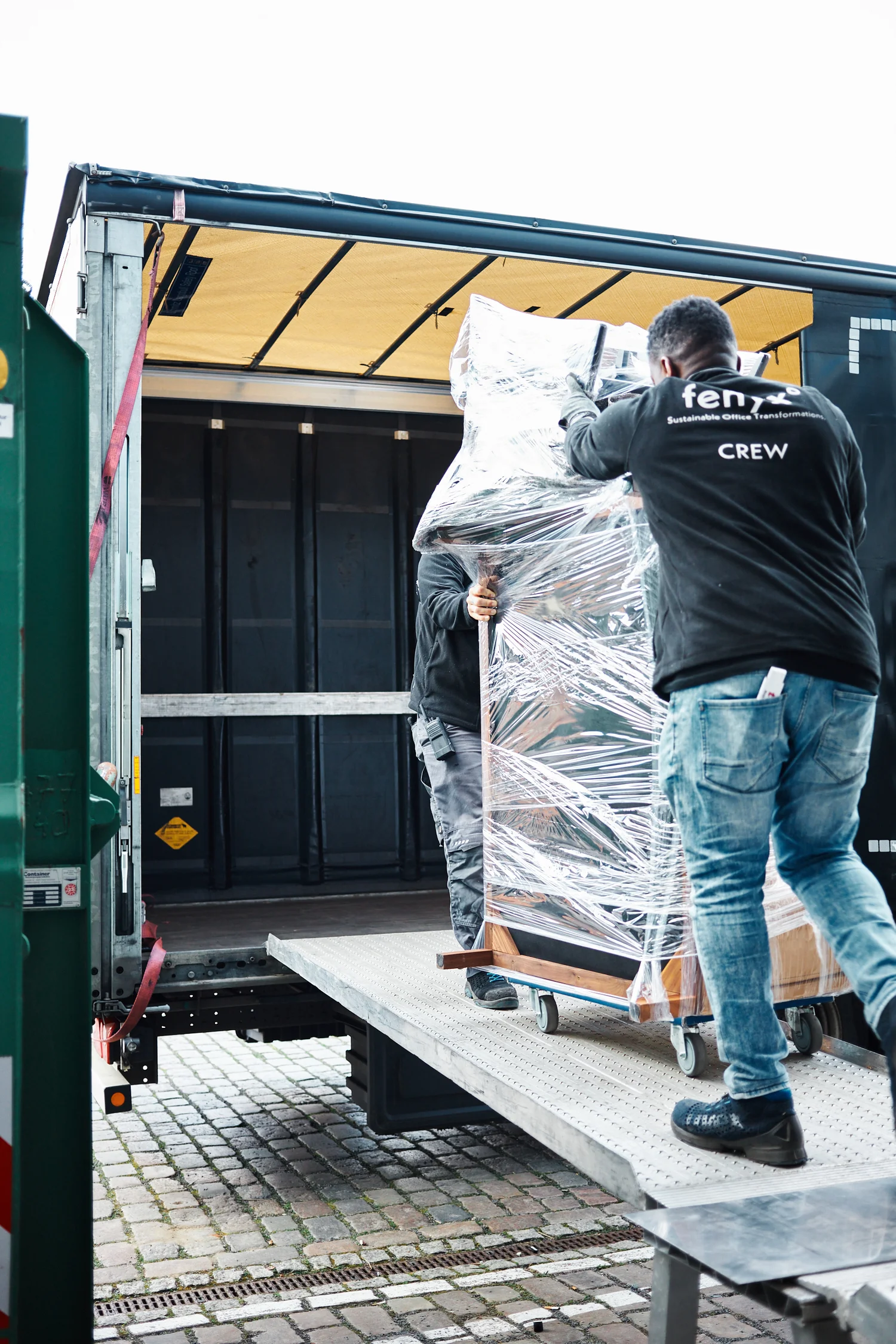 Two crew members loading a large wrapped item on wheels into a truck using a metal ramp over cobblestone pavement.