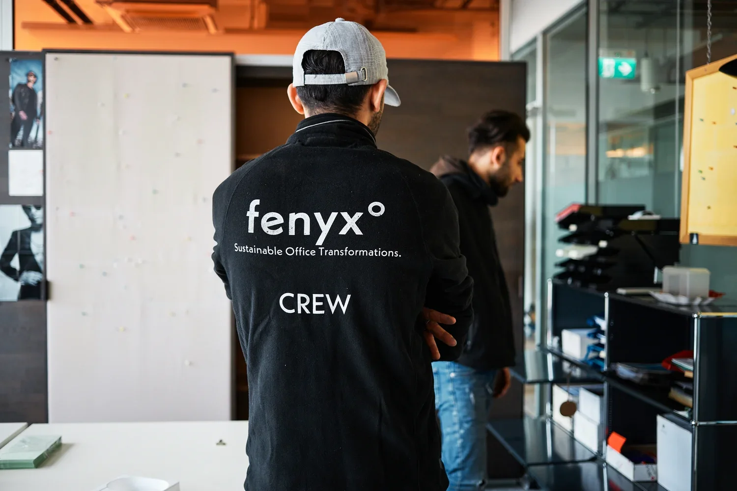 Man wearing a black jacket with 'fenyx Sustainable Office Transformations CREW' printed on the back standing in an office setting.