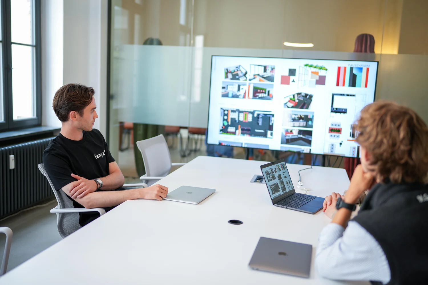 Two men sitting at a white conference table looking at a large screen displaying architectural floor plans and design layouts.