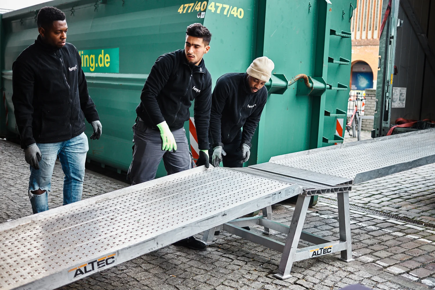 Three workers wearing black jackets and gloves install metal ramps on a cobblestone surface next to a large green container.
