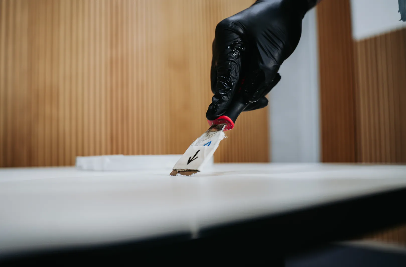Hand in black glove scraping white surface with a putty knife.