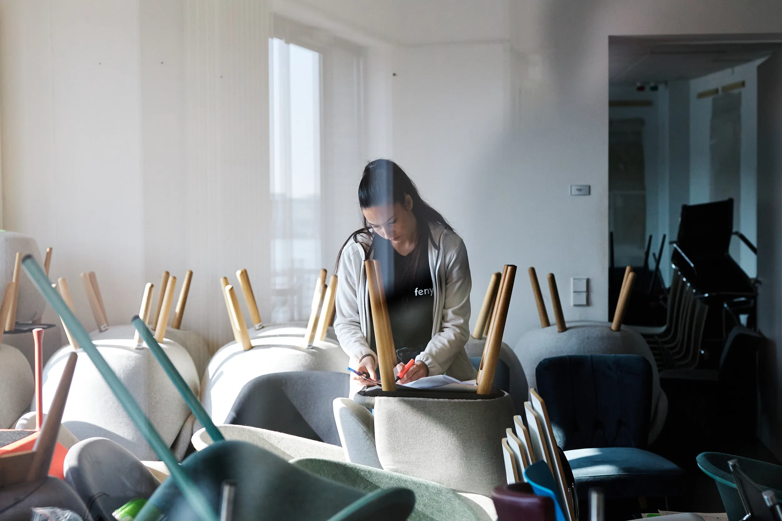 Woman writing on papers while sitting among stacked chairs with wooden legs pointed upward in a bright room.