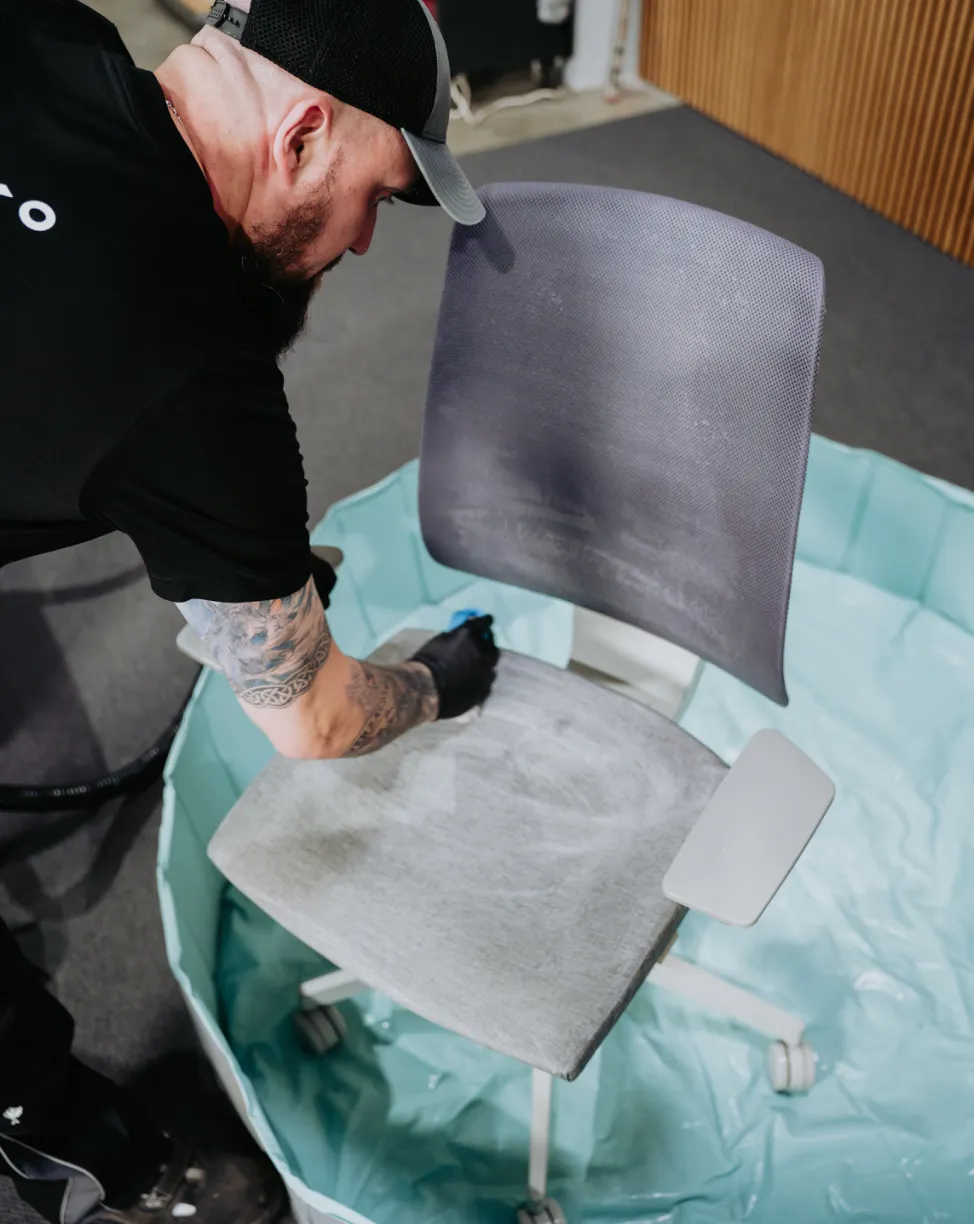 Man wearing a black cap and gloves cleaning a gray office chair inside a blue plastic basin.