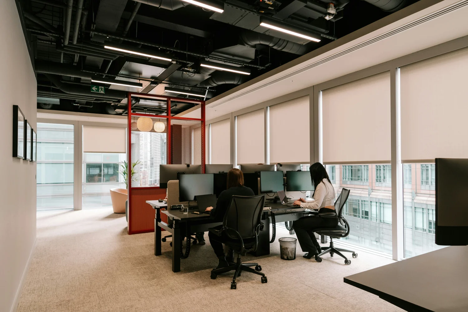 Modern office space with two people working at desks equipped with multiple monitors and rollershade windows.