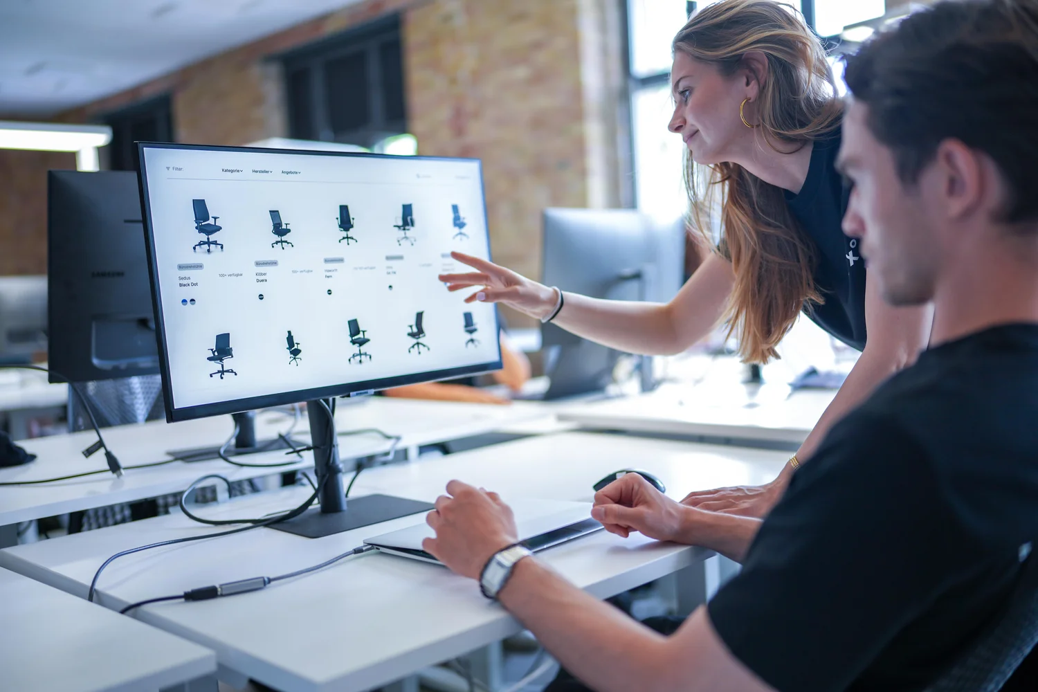Two young professionals collaborating while browsing office chair options on a computer screen in a modern workspace.