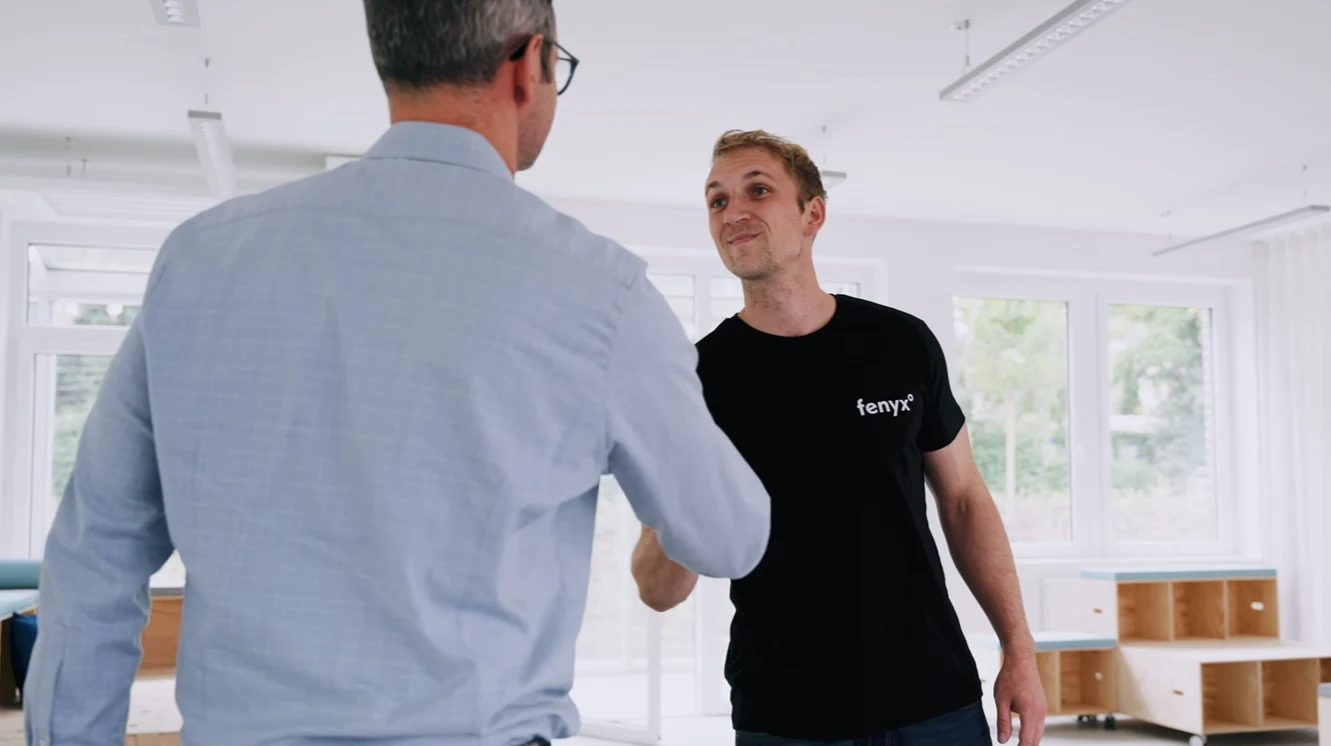 Two men shaking hands in a bright office room, one wearing a black fenyx shirt and the other a blue shirt with glasses.