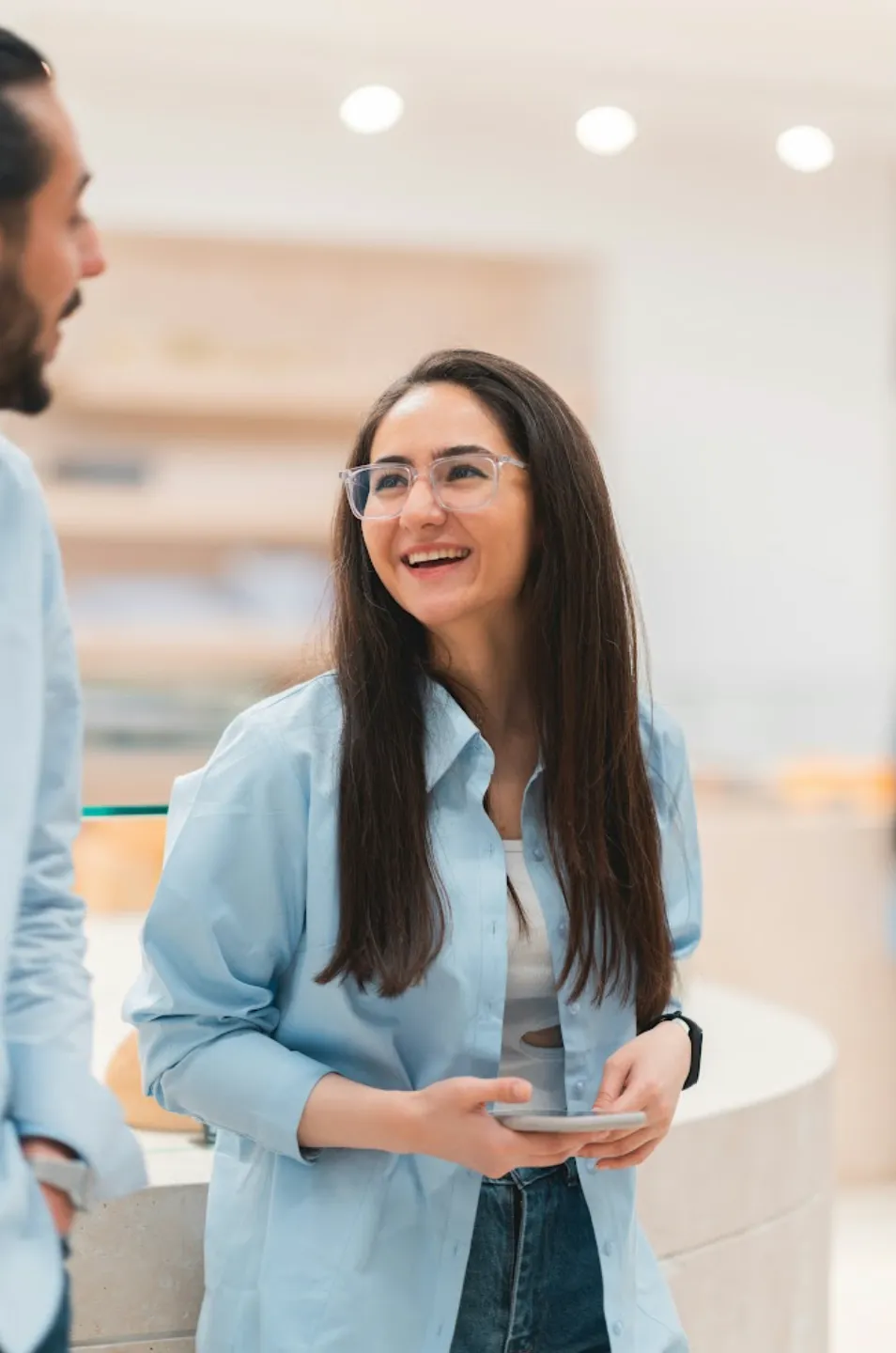 Smiling woman with long dark hair and glasses holding a smartphone, talking with a man.