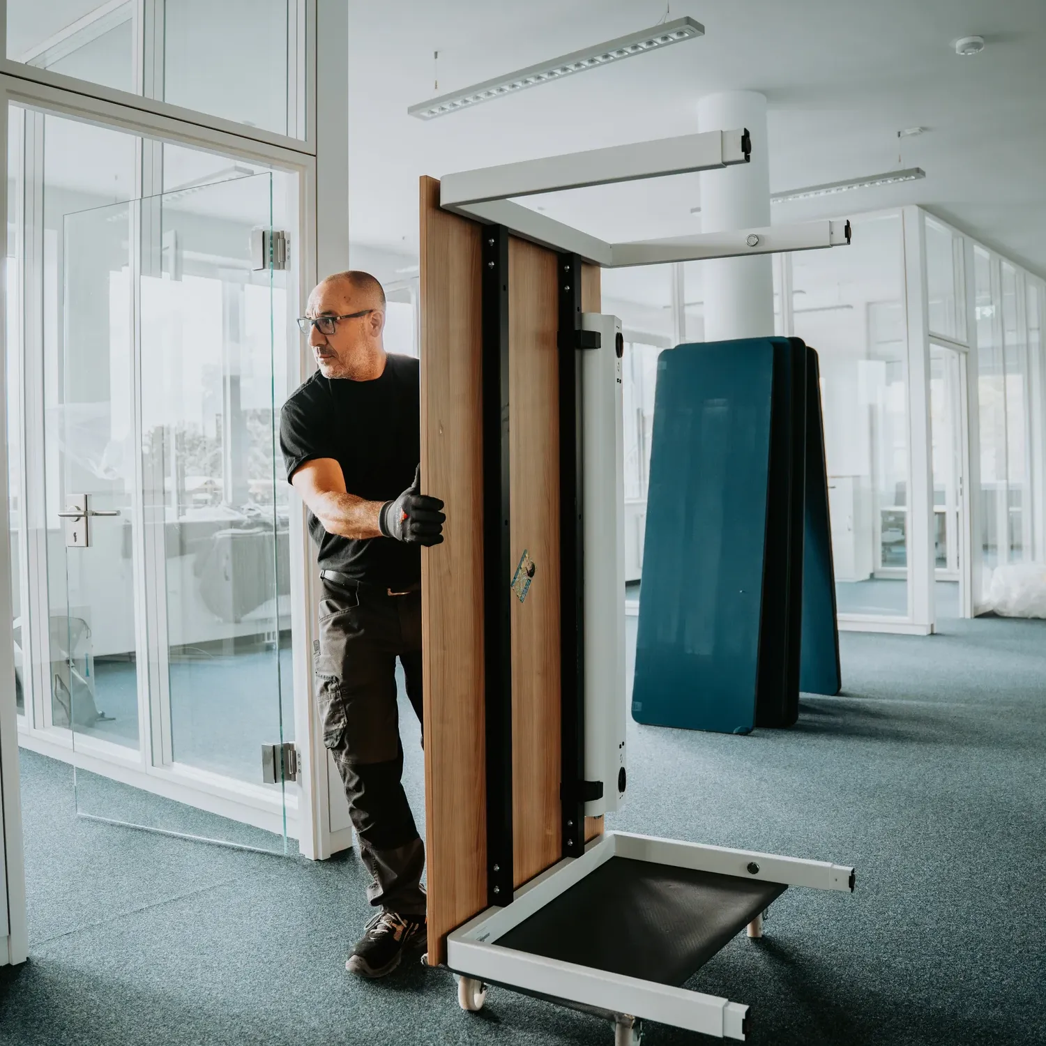 Man wearing gloves moving a folded treadmill inside a modern office space with glass walls.