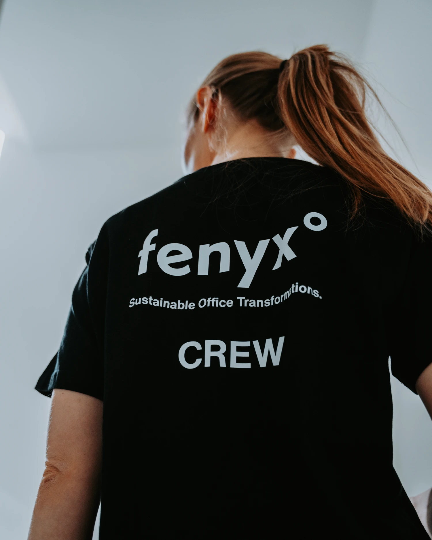 Person with brown ponytail wearing a black shirt with white text reading 'fenyx sustainable Office Transformations CREW' on the back.