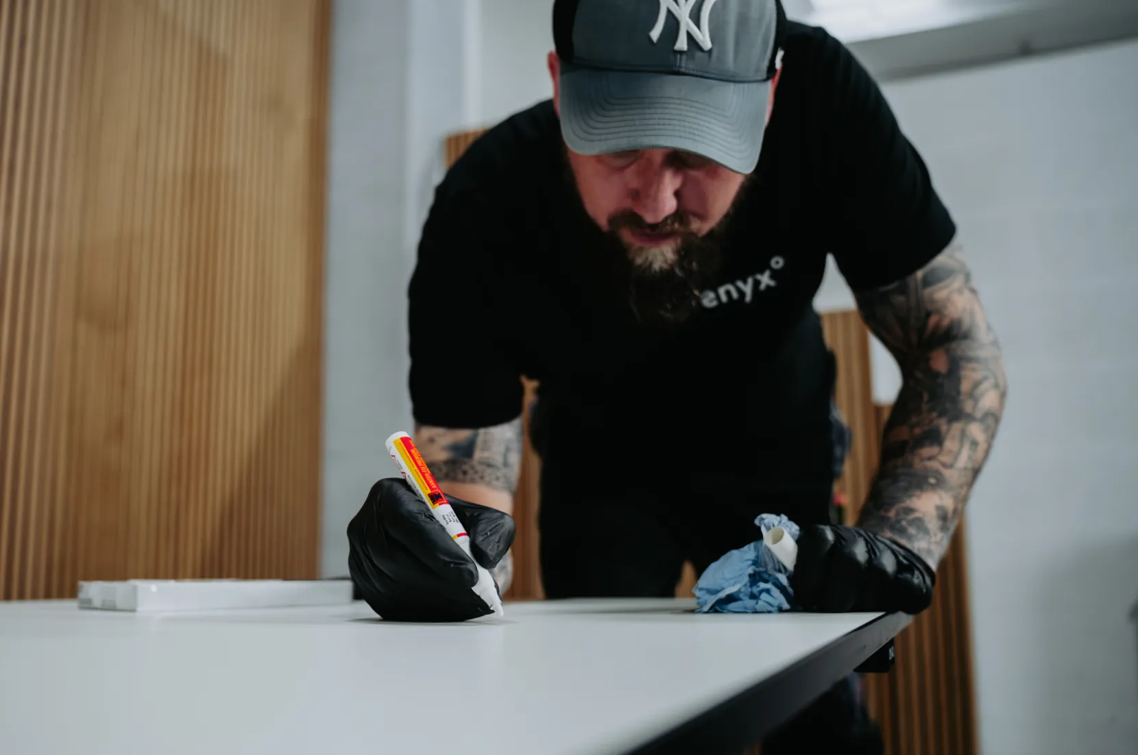 Man wearing tattoos and black gloves applying adhesive on a white surface while holding a blue cloth.