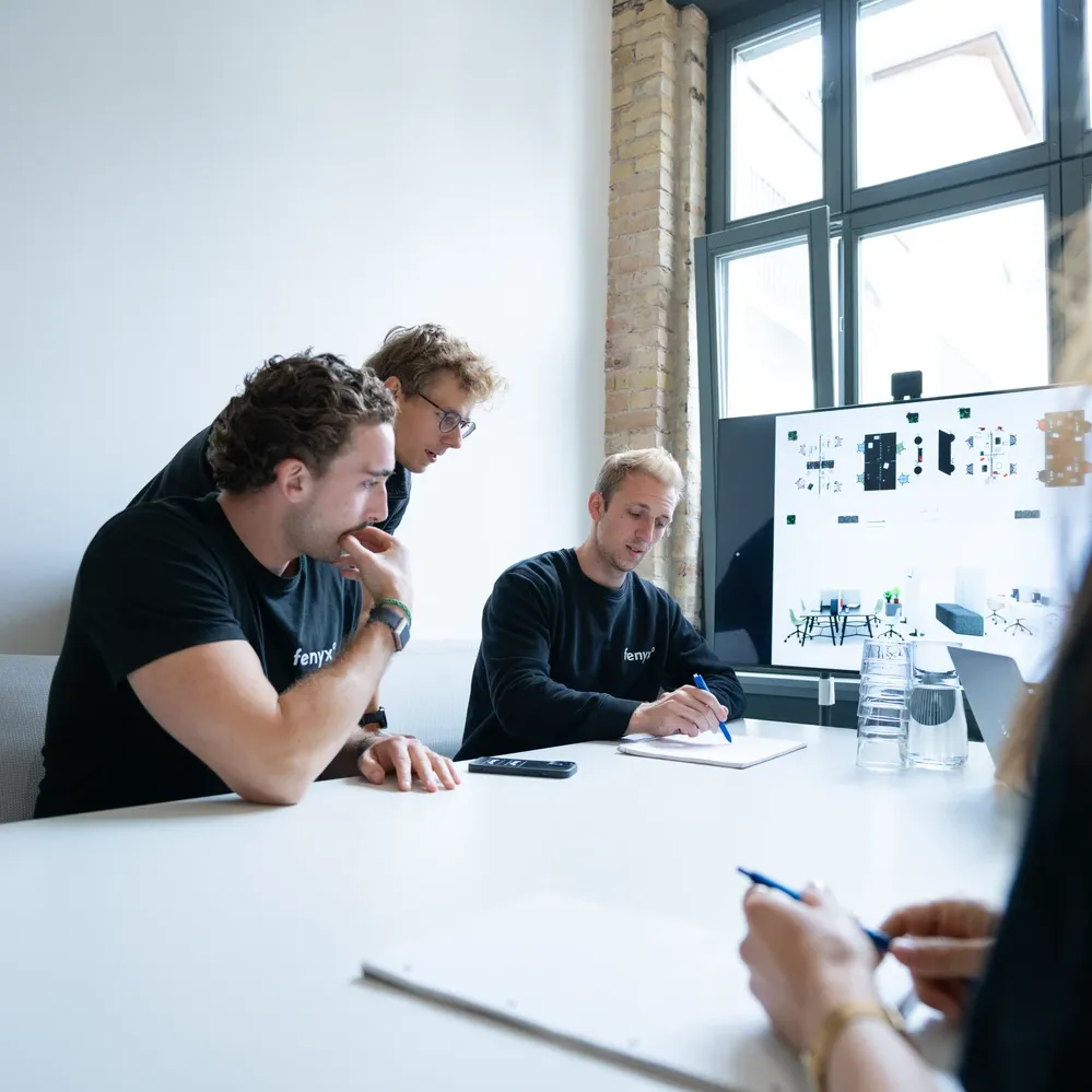 Three men collaborating at a table in a bright office, with one writing on a notepad and a screen displaying diagrams in the background.