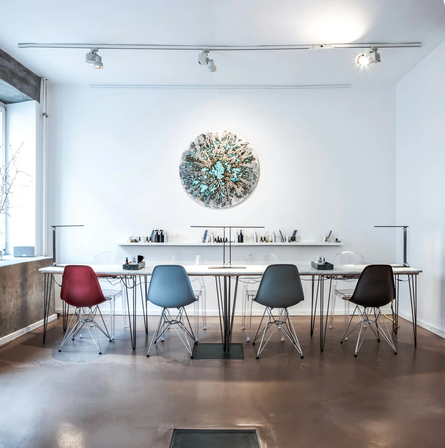 Modern minimalist office with a long table, four colored chairs, clear chairs, and a round abstract wall art above.