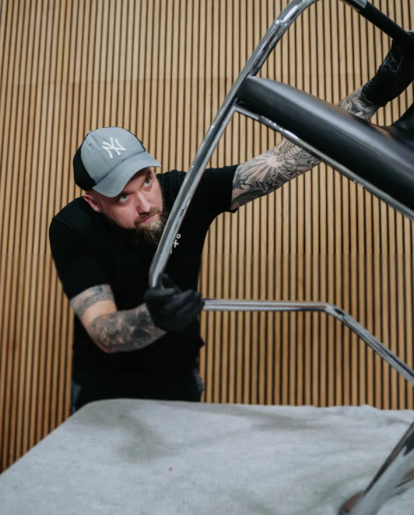 Tattooed man wearing a cap and gloves assembling or adjusting a metal chair frame over a gray surface with a wooden slat wall background.