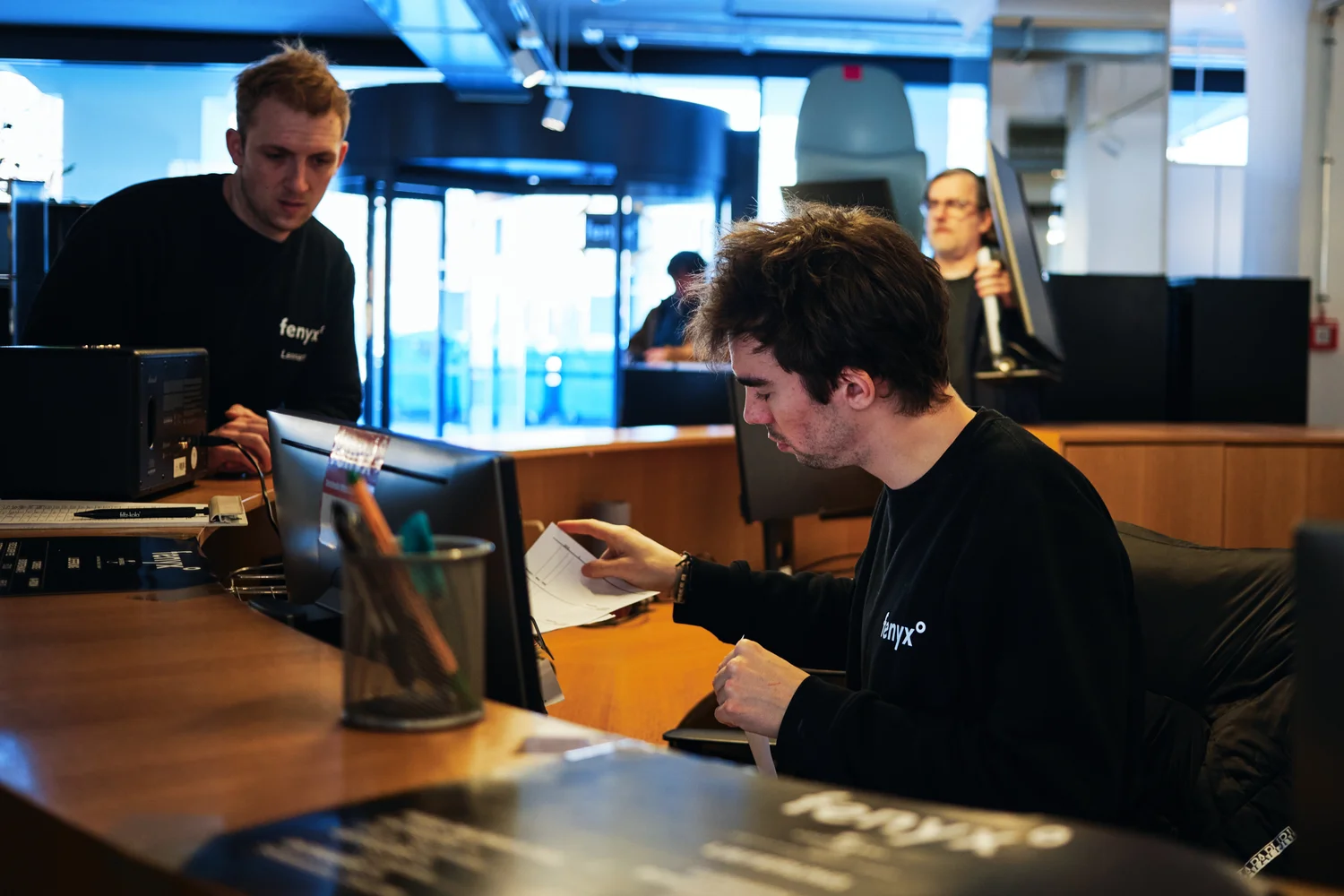 Two men working at a wooden reception desk with computers and papers in a modern office space.