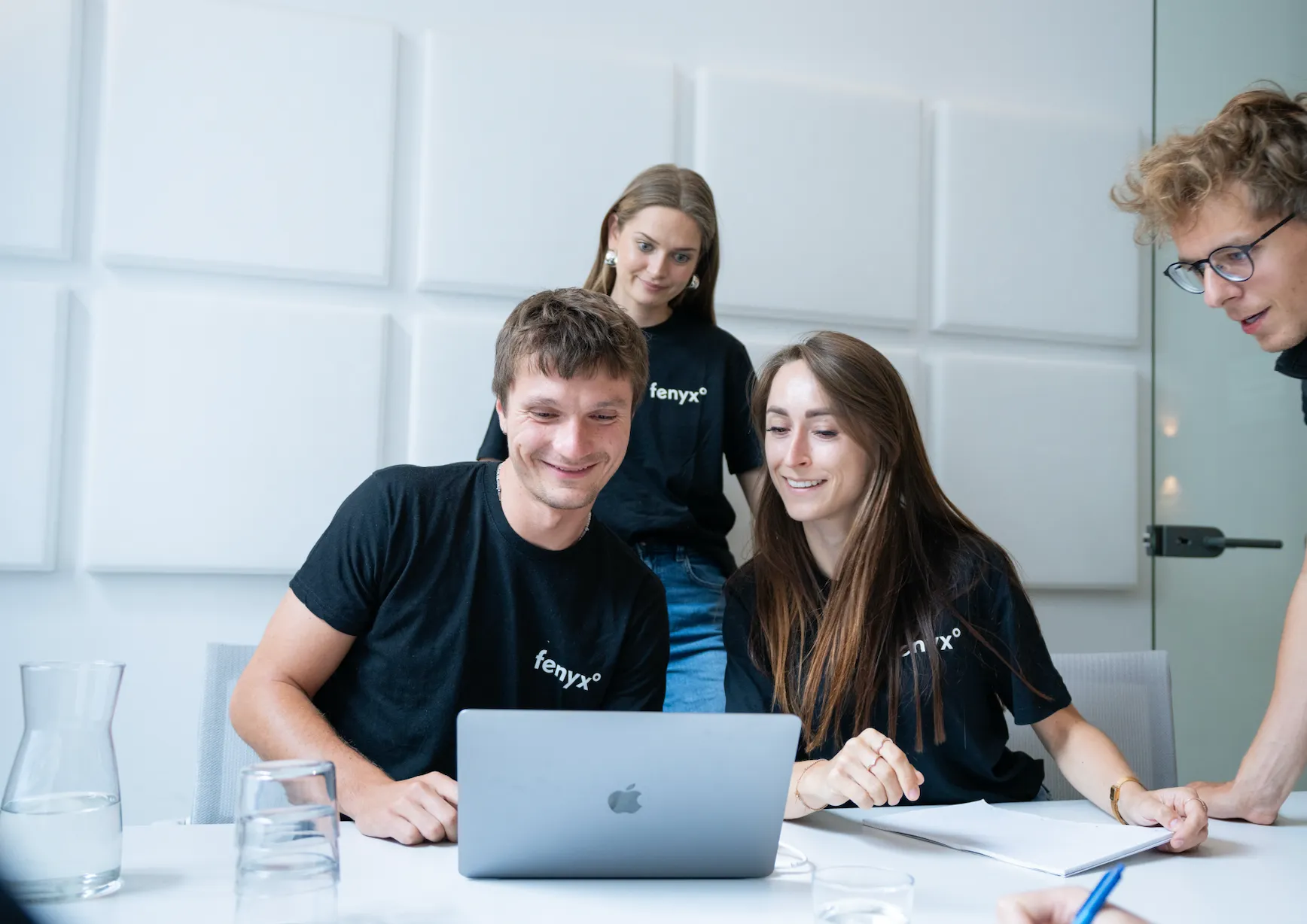 Four young adults wearing black 'fenyx°' shirts collaborating around a laptop in a modern office.