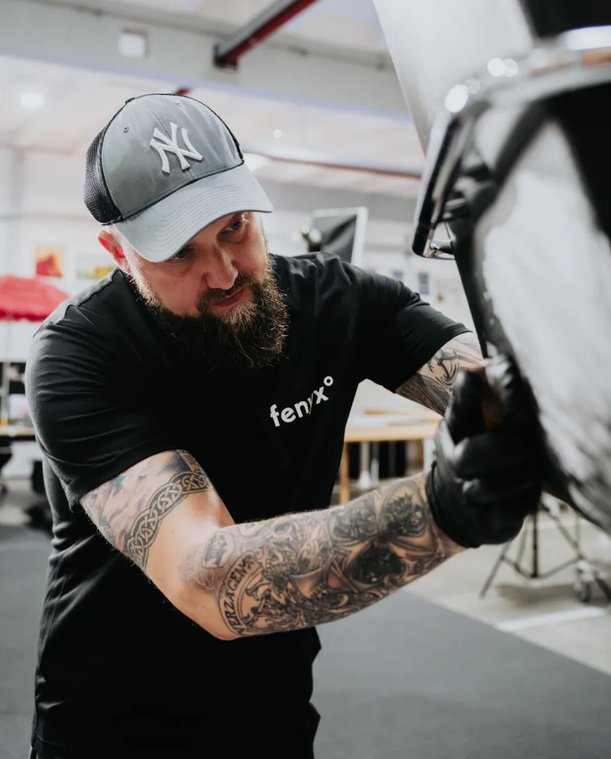 Bearded man wearing a New York Yankees cap and black fenixo t-shirt, working with gloves on a vehicle part in a workshop.