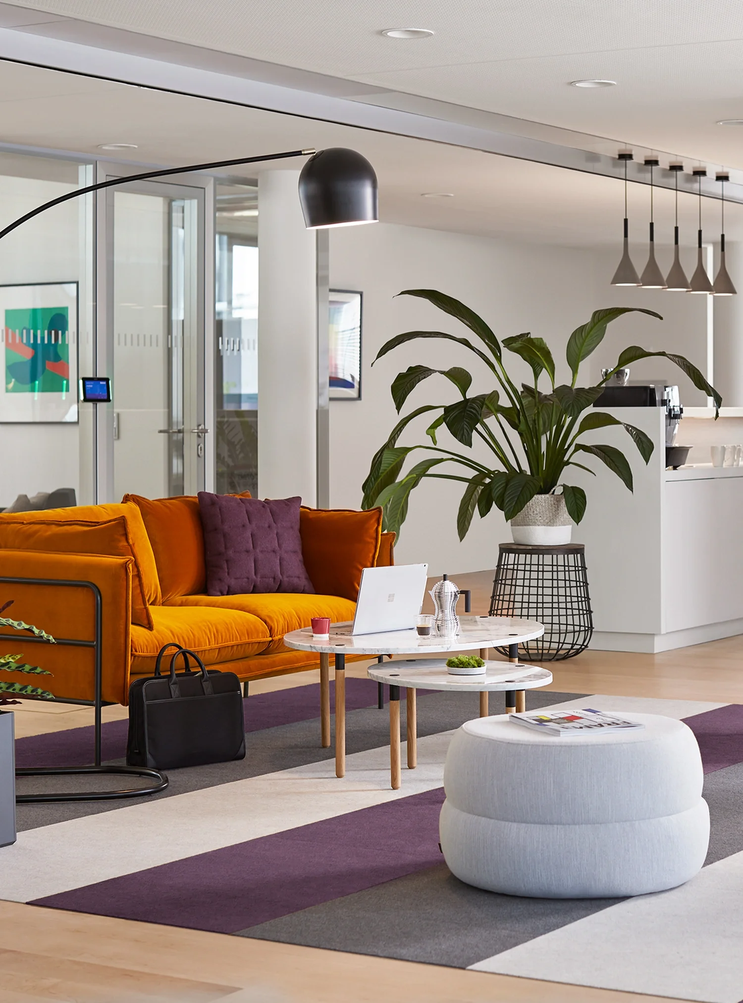 Modern office lounge with orange sofa, purple pillow, round white coffee tables, large green plant, and white pouf on a colorful striped rug.