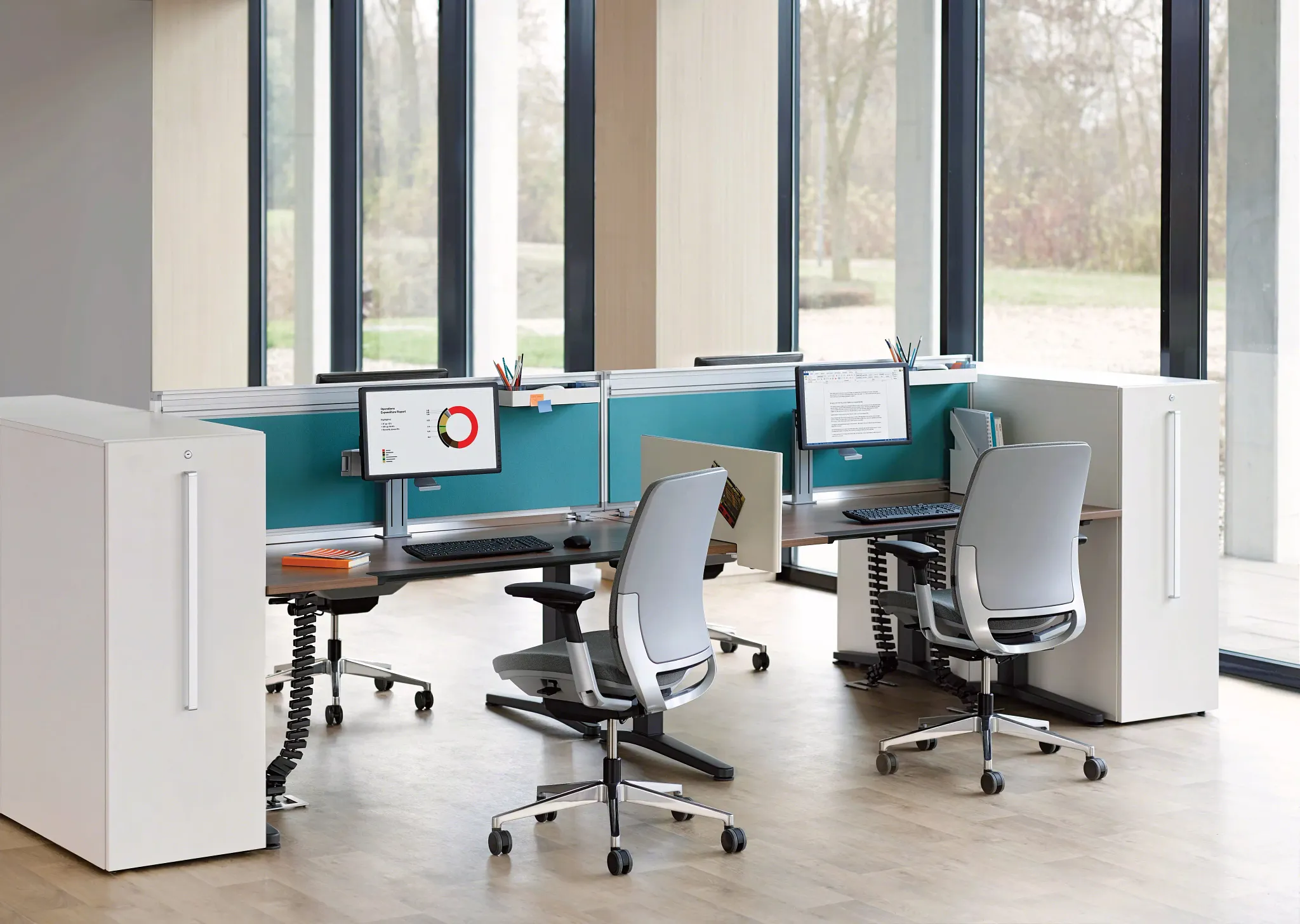 Modern office workspace with two ergonomic chairs, dual computer monitors, and large windows offering natural light.
