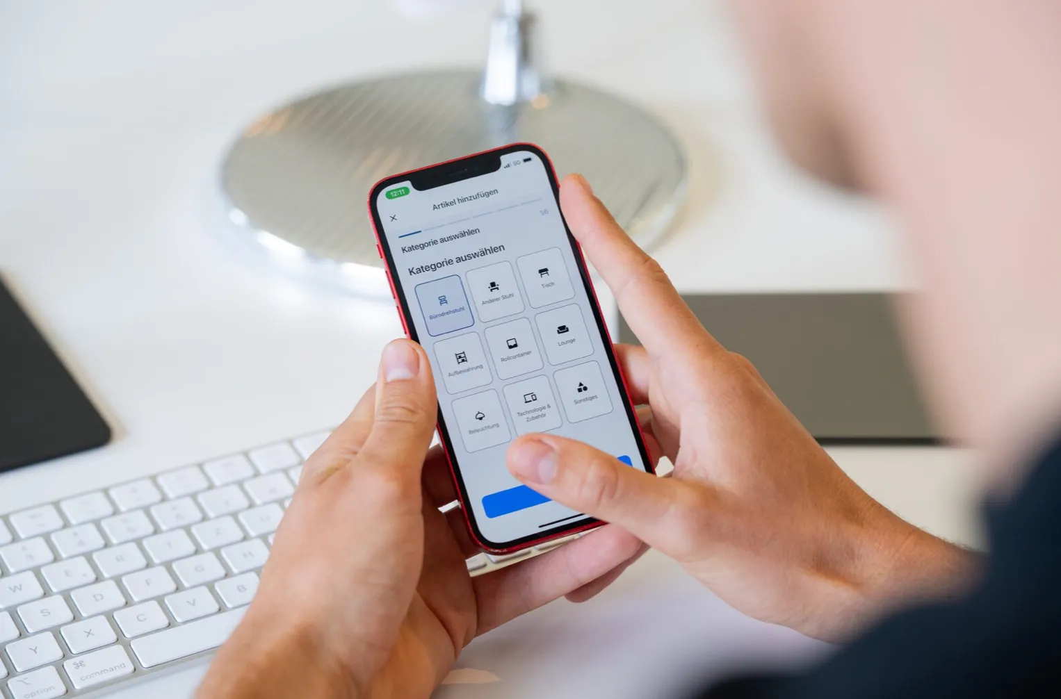 Person holding a red smartphone displaying a German app screen with categories like office chair, other chairs, table, and lighting.