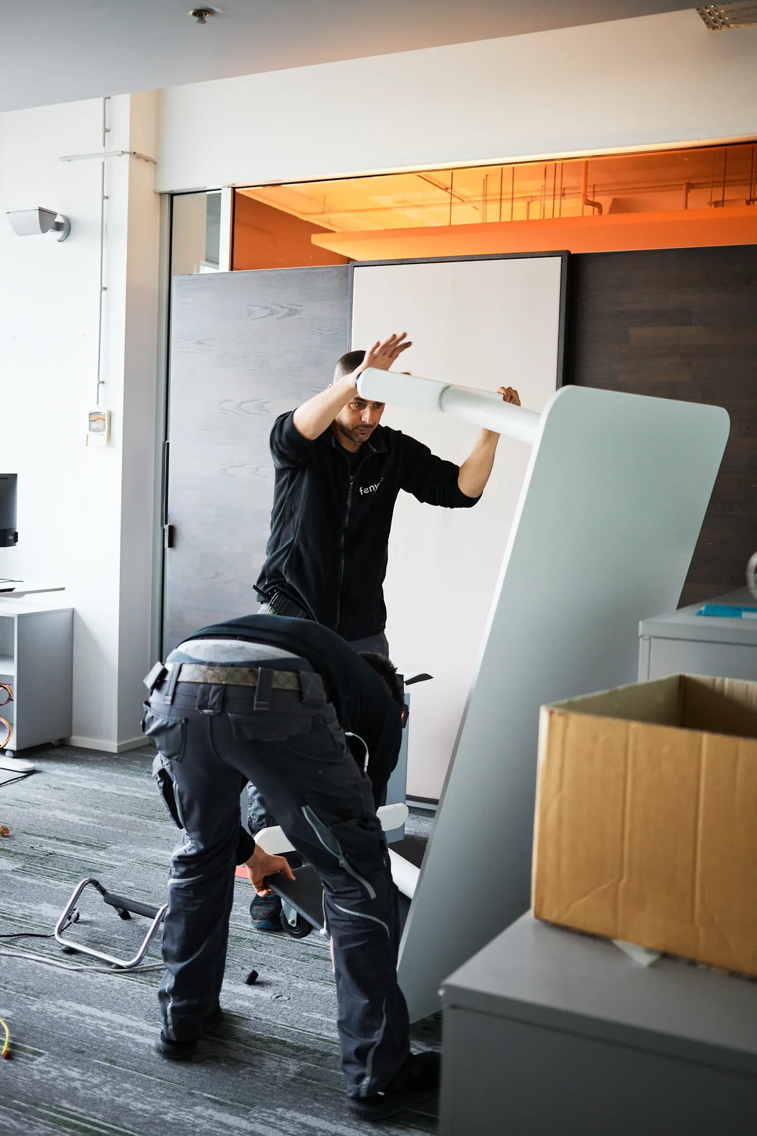 Two men assembling or moving a large whiteboard or panel in a modern office space.