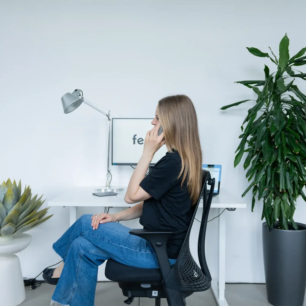 Woman sitting on an office chair, talking on a phone with a desk, computer monitors, and plants in the background.