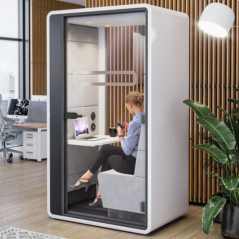 Woman sitting inside a modern office phone booth working on a laptop and checking her phone.