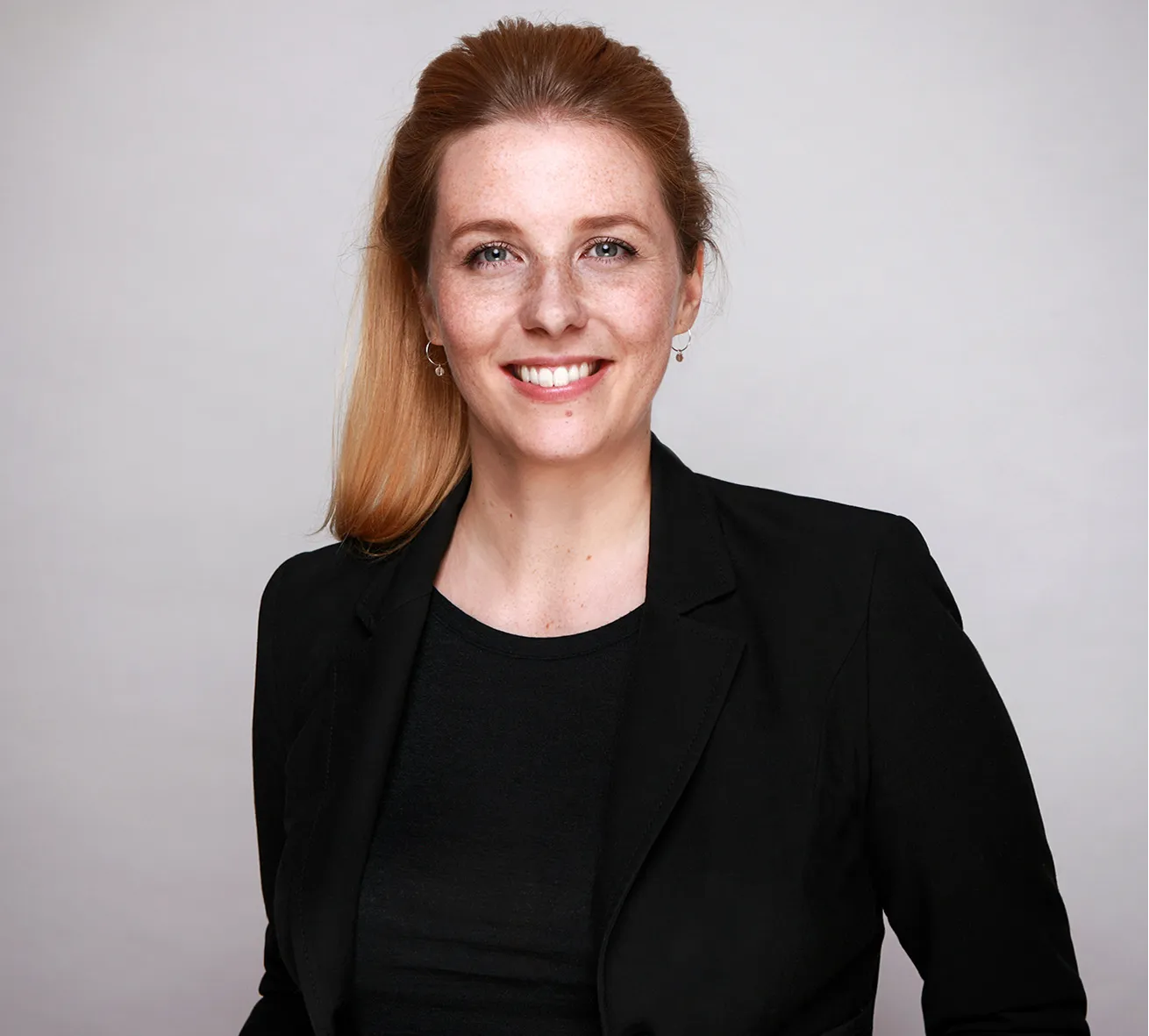 Smiling woman with light red hair, wearing a black blazer and black top, against a plain light gray background.