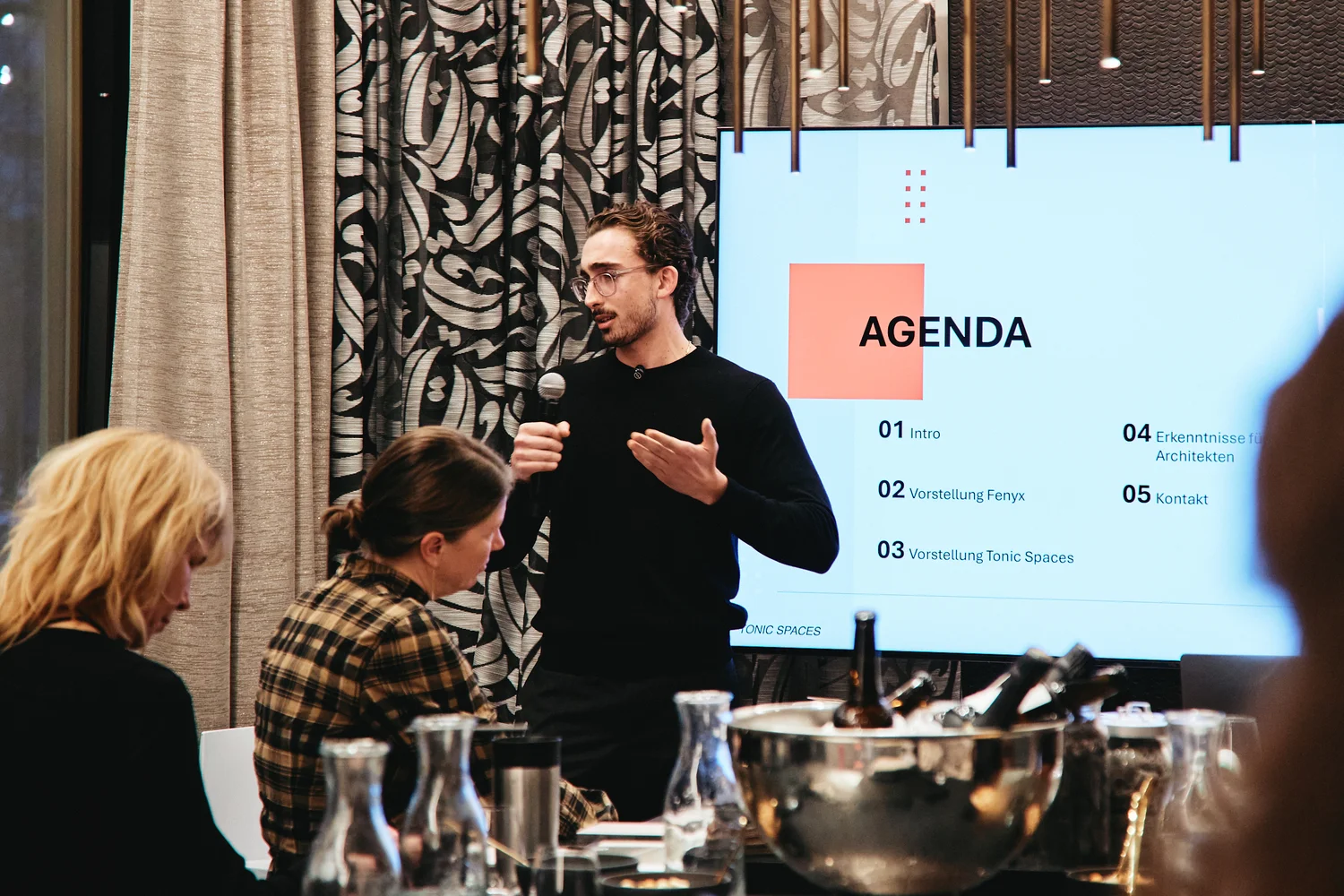 Man in black sweater giving a presentation with a microphone in front of a screen displaying an agenda while two women sit listening.
