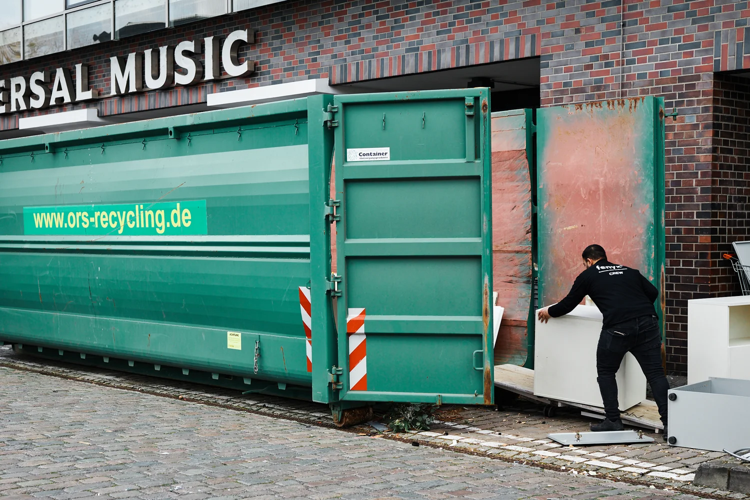 Person loading white furniture into a large green recycling container outside a brick building.
