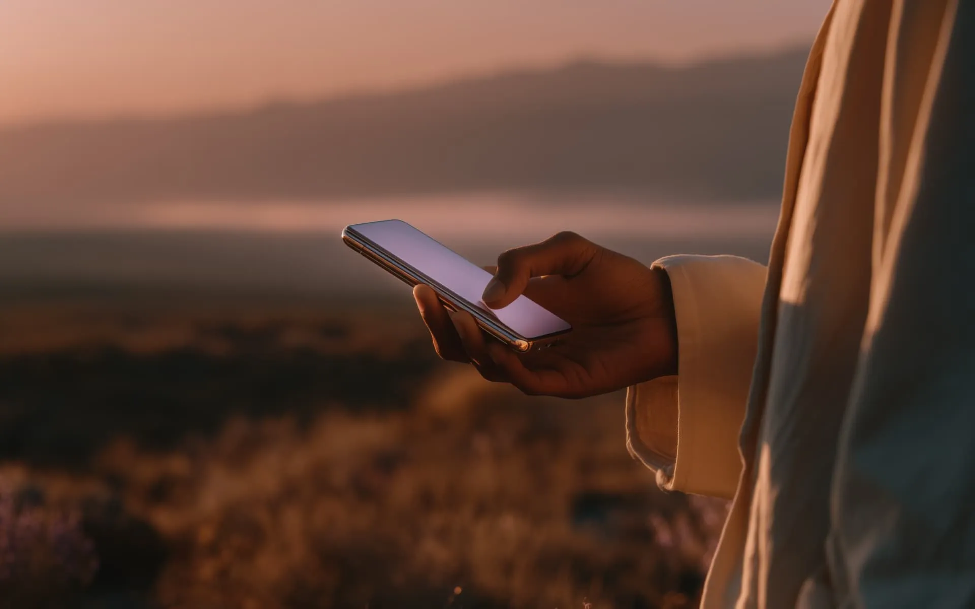 Person holding and using a smartphone outdoors with a blurred natural landscape at sunset in the background.